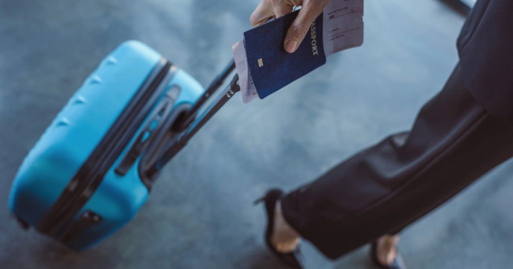 A close-up of a hand holding a passport and ticket, with a suitcase in tow, illustrating travel with cremated remains.