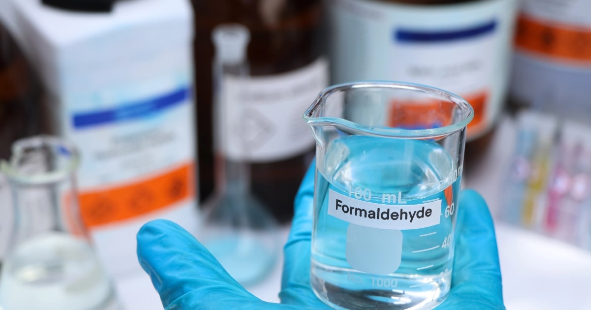 Gloved hand holding a beaker labelled formaldehyde, one of the main chemicals used in embalming.