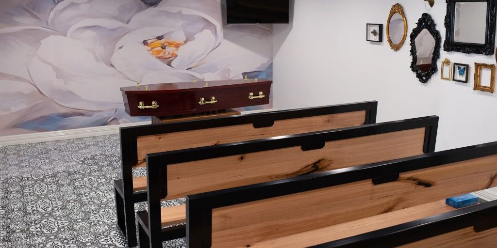 What Is Cremation? - A serene viewing room with unique wooden and black metal benches, a flower wall mural, decorative frames, and a wooden coffin, creating an inviting atmosphere for a memorial.